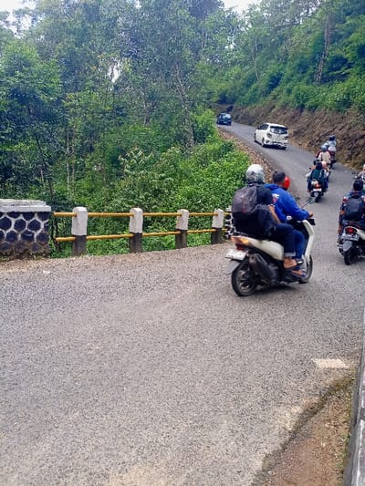 The image shows a scene on a paved road in the Cisewu area of Indonesia, with a bridge crossing a small ravine. The road curves around a hillside covered in dense vegetation. There are multiple people riding motorbikes. Two motorbikes are in the foreground, one with a passenger, heading toward the camera, and the other with a single rider heading away from the camera. A white car is visible going up the hill in the background. The road has a yellow and white guardrail on the right side and the bridge is surrounded by a low wall. The sky is partially visible and appears overcast. The environment is rural and mountainous, with lush green foliage.