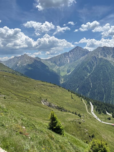 This is a landscape photograph taken in the mountains of St. Leonhard in Passeier - San Leonardo in Passiria, Italy. The image depicts a winding road with several cars and a bus, snaking its way up a lush green mountain slope. The road is bordered by dense coniferous trees on one side and a grassy slope with scattered small trees and rocks on the other. In the background, majestic, rugged mountain peaks rise against a bright blue sky dotted with fluffy white clouds. The overall scene conveys a sense of natural beauty and the vastness of the mountainous terrain.