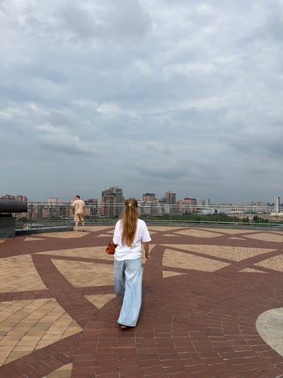 In Tarlashi, Russia, a woman with long blonde hair, wearing a white t-shirt and wide-leg jeans, walks away from the camera on a patterned brick plaza. She carries a brown handbag. To her left, a man in a light tan t-shirt and shorts stands facing away from the camera, looking towards a city skyline. The background shows a dense collection of buildings under a cloudy sky. A metal railing separates the plaza from an area with trees and more buildings in the distance.
