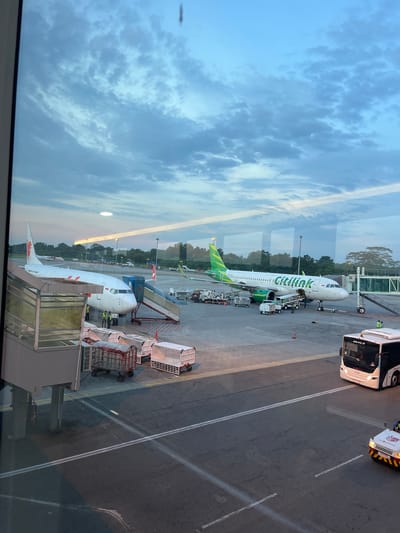 The image, taken from behind a glass window, depicts an airport scene in Ramunia Dua, Indonesia. Two aircraft are visible: one is a white and green Citilink airplane being serviced on the tarmac, with a passenger bus and other support vehicles nearby. The second aircraft is white with a red lion logo, a boarding bridge connected. Airport personnel are visible in hi-vis vests attending to the aircraft. The foreground shows elements of the airport terminal or boarding area behind the glass. The sky is cloudy with hints of sunset colors. The overall environment appears to be a busy airport during the day.