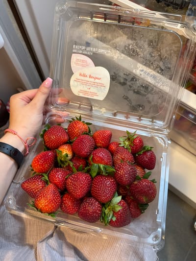 A person's left hand is holding open a clear plastic container filled with ripe strawberries. The container has a hinged lid. The person's fingers are visible around the edge of the container, and their fingernails are painted pink. A black fitness tracker is visible on their wrist, with a pink braided bracelet. The strawberries are a deep red color with green leaves. The background is blurry, but appears to be an indoor setting, possibly a kitchen, with white cabinetry and shelving. The image was taken in the City of Yonkers, United States of America.