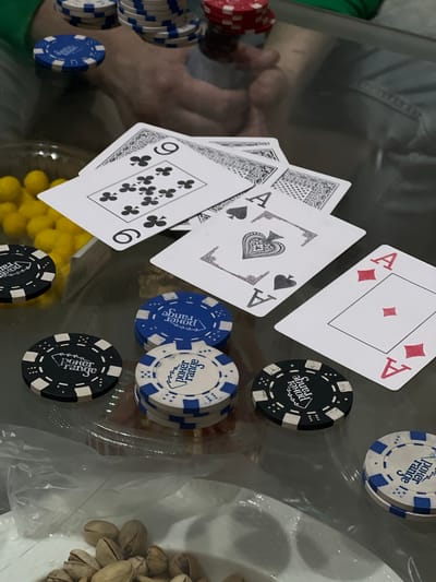 A poker game is in progress on a glass surface. The cards visible include three aces (diamonds, clubs, spades), a nine of clubs, and other partially obscured cards. There are stacks of poker chips of various colors (blue, white, red, black) scattered around the cards. A clear plastic bag filled with yellow candies is partially visible. Pistachios are on a white plate. A person's reflection is visible in the glass surface. There is no indication of the location being Svkh Voronezhskiy, Russia.