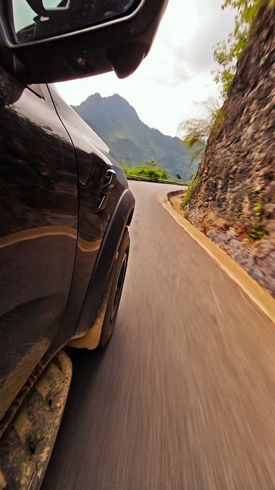 The image captures a moving vehicle, likely a car, on a winding road through a mountainous region. The road curves sharply to the right. A close-up view of the vehicle's side, wheel, and side mirror are visible. The landscape beyond the road features a rugged cliff face on one side and a valley with mountains in the distance. The photograph evokes a sense of motion, with the blur indicating speed. Based on the file information, the location is Cán Tỷ Commune, Vietnam.
