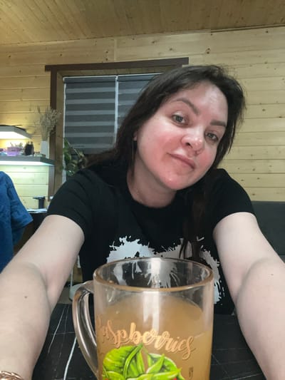 A woman with dark hair and fair skin is posing for a selfie. She is wearing a black t-shirt with a white graphic. Her arms are visible, with a glass mug decorated with a botanical motif near her right arm. The background features wooden walls, possibly indicative of a log cabin or a room with wood paneling. A window with striped blinds is also visible. The location is unknown.