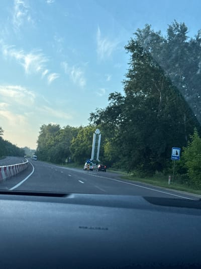 The image depicts a view from a vehicle on a highway, likely in or near the city of Cojușna, Moldova. The road is paved, with white lane markings and a concrete barrier with red and white reflective markings on the left side, suggesting ongoing construction or maintenance. Several vehicles are visible on the road, including a white van further ahead and two cars closer to the foreground on the right lane.
On the right side of the highway, there is a tall, white, monument-like structure with green accents. Adjacent to it, a blue and white road sign with a silhouette of a tree and an arrow pointing to the right is visible. The surrounding environment is lush and green, dominated by dense trees and foliage. The sky is a clear blue with wispy white clouds. The image is taken during daylight, possibly late afternoon or early evening, given the soft lighting. The foreground shows the dashboard of the vehicle from which the photo is taken.