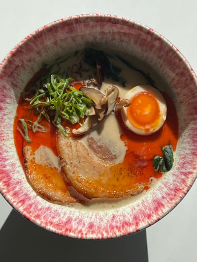 The image displays an overhead, close-up view of a bowl of ramen placed on a plain white surface. The ceramic bowl has a distinctive rim featuring a speckled pattern of white, pink, and dark red hues. Inside the bowl, a creamy, off-white broth is visible, with significant patches of vibrant red chili oil mixed in and floating on its surface, particularly on the left and lower-right portions.
Key components of the ramen include:
- At least two slices of seared or roasted pork belly (chashu) with light brown, slightly caramelized edges, positioned in the lower-center and lower-left.
- One half of a soft-boiled egg (ajitama) located in the upper-right quadrant, revealing a firm white exterior and a bright orange, semi-liquid yolk. A small, dark brown mushroom piece rests on the egg.
- Several slices of dark brown mushrooms are scattered in the upper-center and upper-left.
- A pile of finely sliced green scallions (negi) provides a green accent in the upper-left.
- Dark green leafy vegetables or seaweed are partially submerged in the broth in the upper-center and lower-right.
The scene is illuminated from the upper-left, casting a distinct dark shadow from the bowl onto the white surface in the lower-left corner. No people, discernible actions, or interactions are visible. The background consists solely of the white surface. The location within the city of Pozuelo de Alarcón, Spain, cannot be determined from the visual information provided in this image.