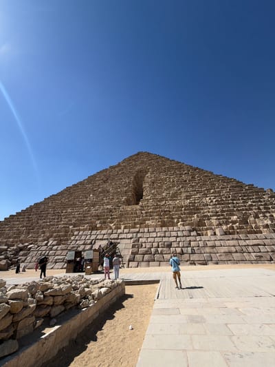 In this vertical, eye-level shot, the massive, rough-hewn stone facade of a pyramid dominates the frame against a clear, bright blue sky. The weathered, tan-colored blocks are stacked in an orderly yet uneven fashion, creating a textured surface that hints at great age. A large, dark rectangular opening interrupts the stonework midway up the pyramid, suggesting an entrance or a void.

Along the base of the pyramid, a paved pathway made of lighter, rectangular stones stretches out. Several people are visible on this path, appearing small in comparison to the monumental structure. One person, wearing a light blue checkered shirt and brown shorts, stands alone, casting a long shadow onto the paved area. Further along the path, a small group of people are gathered, some appearing to interact with what might be information booths or ticket stands, indicated by the presence of signs with visible text. The ground bordering the paved area is sandy and uneven, scattered with rocks.

The strong sunlight casting sharp shadows indicates a time of day when the sun is high, likely midday. The clear sky suggests fair weather. The overall scene evokes a sense of awe at the scale of the ancient structure and the bustling activity of tourists exploring the historic site. The location is identified as Kafrat Nassar, Egypt, implying this is likely a well-known pyramid complex.