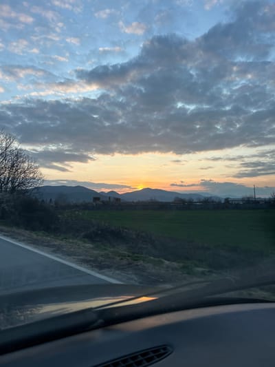The image depicts a rural sunset scene in Vedrare, Bulgaria, viewed from inside a vehicle. The sky occupies the upper two-thirds of the frame, featuring a gradient from light blue at the zenith to bright orange and yellow at the horizon where the sun is visible as a distinct orange disc just above a distant, undulating mountain range. Overcast cloud cover is present, with grey and white stratocumulus clouds scattered across the sky, particularly dense in the upper center and right.
In the mid-ground, a broad, vibrant green field stretches horizontally across the scene, bordered by dark, uncultivated ground and sparse vegetation in the immediate foreground. To the left of the field, several bare tree branches protrude into the frame. Along the distant edge of the field, situated against the backdrop of the mountains, are scattered structures. On the left, a cluster of dilapidated, dark brown buildings with partial roofs is visible. To the right, further structures appear, including indistinct low-rise buildings and utility poles with power lines extending towards the right edge of the frame.
The foreground is dominated by the dark grey dashboard and lower windshield of a vehicle. A portion of asphalt road with a white lane marking is visible through the windshield on the lower left. A reflection of the setting sun is observable on the dashboard or windshield. No individuals are visible in the scene.