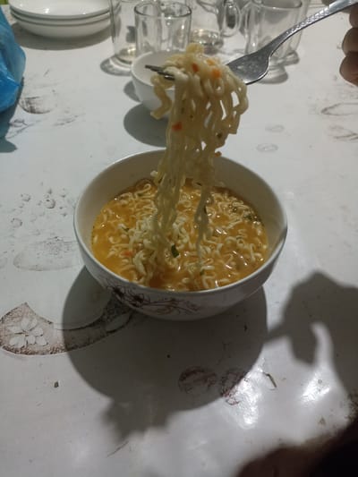 A bowl of ramen noodles is pictured on a patterned tablecloth. A fork is lifting a portion of the noodles out of the bowl. In the background, there are several glasses and a blue plastic bag. The scene appears to be indoors, possibly in a home setting in Кош-Дөбө, Kyrgyzstan. The lighting suggests it may be evening or a dimly lit room, casting shadows from the bowl and fork onto the tablecloth. The focus is on the food, with the background elements slightly blurred.