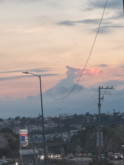 The image depicts a scenic view of a mountain, likely Popocatépetl, emerging from a layer of clouds at sunset. The sky is a gradient of soft pinks and blues, with wispy clouds adding texture. In the foreground, a street light and power lines are visible, along with a Mobil gas station sign displaying prices. The scene also includes a highway with several cars, suggesting this is a view from the outskirts of a city. The city of San Francisco Totimehuacán, Mexico, is known for its proximity to active volcanoes and its developing urban landscape, which aligns with the visual elements presented in the image. The overall atmosphere is tranquil, capturing the natural beauty of the region under the soft glow of the setting sun.