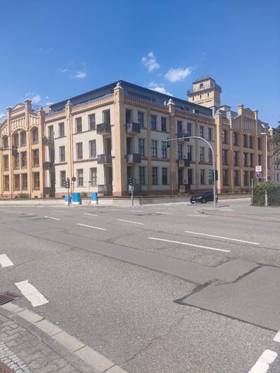 The image depicts a bright, sunny day in Chemnitz, Germany, with a clear blue sky dotted with a few wispy clouds. The scene is dominated by a large, modern apartment building with a distinctive architectural style that blends industrial elements with contemporary design. The building features a pale yellow facade with accents of red brick and numerous windows. Several balconies with dark metal railings punctuate the exterior. A prominent tower with a pitched roof rises from one section of the building, adding a unique character to the structure.
In front of the building, a multi-lane paved road is visible, marked with white lane lines and a pedestrian crossing. Two black cars are parked on the side of the road. Traffic lights and street signs are present at an intersection, indicating a functional urban environment. Several blue, rectangular utility boxes are positioned along the sidewalk in front of the building. The overall impression is one of a well-maintained and active urban area on a pleasant day. No people are clearly visible in the image. The time of day appears to be late morning or early afternoon, judging by the sunlight and the shadows cast. A "30" speed limit sign is visible on a pole.
