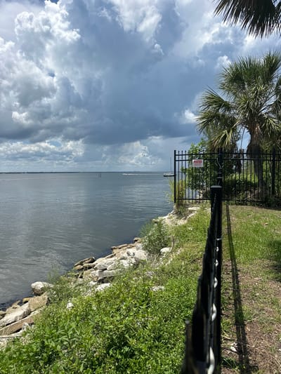 The image shows a body of water with a cloudy sky. A black metal fence runs along the right side of the frame, with a red sign that says "POSTED" attached to it. Palm trees and green vegetation are visible behind the fence. In the distance, there are boats on the water and land on the horizon. The scene appears to be a waterfront area, possibly a park or public access point, in Melbourne, Florida, United States of America. The water is calm, and the clouds suggest an overcast or potentially stormy day. The fence and sign indicate restricted access to the area behind them.