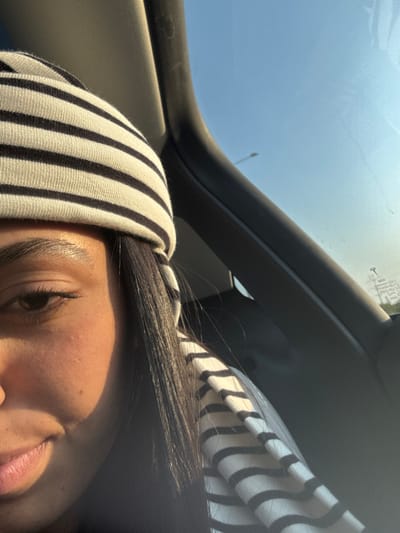 The image depicts a close-up, partial view of a person, likely a passenger, inside a vehicle in Kefar Sava, Israel. The subject, appearing female, is seen from the left eyebrow down to the lower lip on the left side of the face. The left eye is partially visible, and a slight upward turn of the mouth suggests a subtle smile. The skin on the visible facial area has some minor blemishes. Long, straight, dark hair extends from beneath a head covering. The head covering is a beanie or similar item with prominent horizontal black and white stripes. Direct sunlight illuminates the visible portion of the face, creating a strong contrast with shadowed areas. The interior of the vehicle is visible, including a dark-colored A-pillar or door frame on the right and part of the headliner at the top. Outside the vehicle, a clear blue sky is visible through a window, with a faint, distant dark pole-like object. Illegible text or reflections are present on the window glass. The lighting indicates daytime conditions.