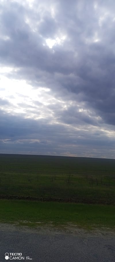 The image shows a landscape in Verkhnyaya Gryaznukha, Russia. In the foreground, there is a gravelly surface, transitioning into a grassy area. Beyond that, a vast green field stretches towards the horizon. Above the field is a sky dominated by heavy, dark, and layered clouds, with patches of sunlight breaking through. There are no people or distinct objects visible in the field. The overall atmosphere is overcast and possibly hinting at rain. In the bottom left corner of the image there is a logo for "Tecno Camon 19 Neo".