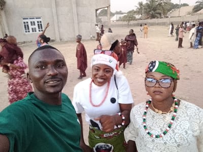 The image depicts an outdoor gathering in Nassarawa Egon, Nigeria. Three individuals are prominently featured in the foreground, facing the camera. On the left, a dark-skinned male with a short haircut and light facial hair wears a dark green short-sleeved shirt and exhibits a slight smile. In the center, a dark-skinned female wears a white short-sleeved shirt, a green patterned wrapper, a red and white beaded necklace, and a red and white Santa-style hat with "Merry Christmas" text, sporting sunglasses pushed onto the hat and a broad smile. On the right, a dark-skinned female wears a white lace top, a multi-colored beaded necklace (red, green, black, white), a gold-colored necklace, large tortoise-shell eyeglasses, and a green, black, white, and red patterned cap, displaying a neutral expression.

In the midground and background, approximately fifteen to twenty additional individuals are scattered across an unpaved, sandy ground, some appearing in traditional Nigerian attire. One person in the midground, to the left, is seen with an arm raised. The background includes a light-colored concrete building with a white-framed window on the left. Further to the right, distant buildings with corrugated metal roofs and sparse vegetation, including palm trees, are visible. The overall scene suggests a social or communal outdoor event under soft natural light.