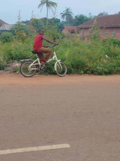 A person rides a white folding bicycle on a dirt road adjacent to a paved road with a white line marking. The rider, wearing a red shirt and brown pants, is surrounded by green foliage. Two houses with red roofs are visible in the background, along with palm trees against a cloudy sky. The scene suggests a rural or suburban environment.