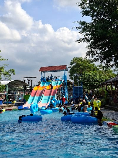 A water park in Kecamatan Tulung, Indonesia, located at coordinates -7.592 latitude and 110.65 longitude. The scene features a large, colorful water slide with people climbing and sliding down, some landing in an inflatable ring. There are many people present, both adults and children, interacting around the slide and pool area. There are life jackets and lifeguards present. Some people are on the slide, some are in the pool, and some are sitting on the edge of the pool or on the ground. The slide is a metal structure with a wooden platform at the top. The pool is a bright blue color and the water is clear. The surrounding vegetation is green and lush. The sky is blue with some white clouds.