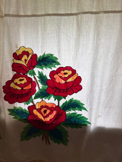 An embroidered floral pattern of four red and yellow roses with green leaves is depicted on a light-colored fabric. The embroidery is detailed, with visible stitches forming the petals and leaves. The fabric appears to be a natural fiber, possibly linen or cotton, with a slightly textured weave. The roses are arranged in a cluster, with stems and leaves extending outwards. The overall impression is of a handcrafted textile, likely a curtain or tablecloth. There are no people or any other objects visible in the image. The background is a plain, light-colored surface, possibly a wall or another piece of fabric. The image has a soft, diffused lighting. The location in the city of Ильпанур, Russia, is not discernible from the image content.
