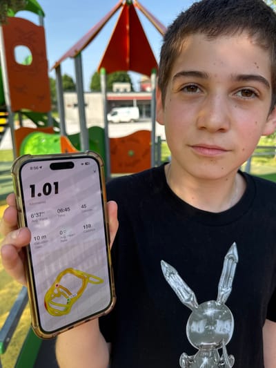 A young boy is holding up a phone that displays a running app showing that he has run 1.01 kilometers in 6 minutes and 45 seconds at an average pace of 6 minutes and 37 seconds per kilometer. He has burned 45 calories. His heart rate is 0 beats per minute and his cadence is 159. The map on the phone shows he has run in a loop that is about 1 kilometer in length. He is wearing a black t-shirt with a silver design. Behind the boy is a brightly colored playground. Based on the presence of playground equipment and the architectural style of the buildings in the background, the image appears to be taken in a public park. It is daytime and the weather appears to be sunny.