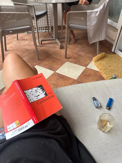 The image depicts an outdoor balcony or terrace in Zadar, Croatia. The foreground features the bare legs of an individual, clad in dark fabric, resting on a light grey upholstered surface. The individual holds an open red paperback book titled "The Collector" by John Fowles, a Vintage Classics edition, its back cover partially visible. Adjacent to the book, on the grey surface, are a small dark grey bottle labeled "Dior Homme," a blue disposable lighter, and an empty, clear, faceted glass. The floor is tiled in a checkerboard pattern of brown and white squares. In the background, a circular grey metal table is positioned among four grey outdoor chairs with perforated seats and backs. The bare feet of at least one other person are visible beneath the table. A white piece of fabric is draped over one of the chairs. To the right, a crumpled yellow, fuzzy rug or blanket lies on the floor, next to a partially visible green bottle with a white cap. A white window or door frame is visible on the far right.