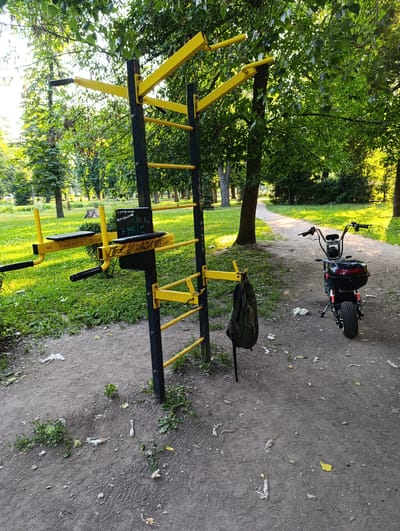 The image shows an outdoor exercise station in a park setting. The station is made of black metal with bright yellow bars and supports, featuring various bars for pull-ups, dips, and other calisthenics exercises. A dark green backpack is hanging from one of the bars.
To the right of the exercise station, a small electric scooter is parked on a dirt path. The scooter is black with red accents and a storage box on the back. The path curves into the background, lined with trees and green grass. Sunlight filters through the lush green leaves of the trees, suggesting it is daytime, likely late afternoon due to the warm light. The overall scene is peaceful and outdoorsy.
There is some visible text on a black board attached to the exercise station. It appears to be graffiti or a message written in white and green markers. Some of the visible text includes what looks like handles or names.
The location is Fastiv, Ukraine. The environment appears to be a public park or recreational area, possibly a sports ground within a larger park. The presence of the scooter might indicate a local resident or someone using it for convenience to reach the park. The weather seems pleasant, with clear skies and foliage indicating a warm season, likely summer.