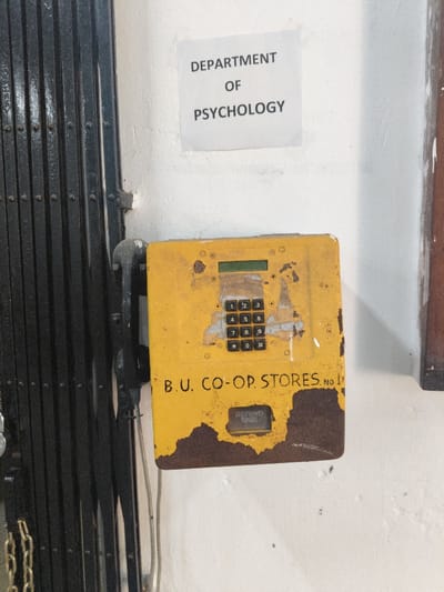 A sign reading "DEPARTMENT OF PSYCHOLOGY" is posted above a weathered, yellow, push-button payphone. The phone is labeled "B.U. CO-OP STORES. NO 1." and has a refund button. The paint is chipping, revealing rust underneath. A black metal security gate is on the left. The wall behind the phone and sign is white.