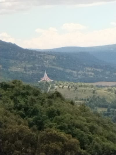 This image presents a wide view of a natural landscape with a prominent structure, located in Waseges ward, Kenya. In the midground, a distinctive building features a multi-tiered, conical roof, patterned with alternating red and white sections, culminating in a slender finial at its apex. This structure is situated on a gentle slope surrounded by green vegetation. The immediate foreground consists of dense, dark green tree canopies, indicating a forested hillside from which the photograph was taken. Beyond the central building, the terrain continues with rolling green hills and valleys, showing varied levels of tree density and patches of lighter, possibly grassy, ground. The distant background reveals multiple layers of blue-tinted mountain ranges receding into the horizon. The sky above is a light blue with scattered white cumulus clouds. No individuals, animals, or explicit actions are visible within the scene.