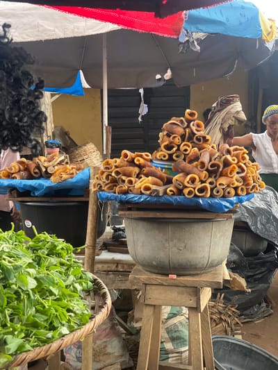 In Ojodu, Nigeria, a street market scene presents a display of processed cow skin, locally known as "ponmo," hung in elongated rolls from wooden beams. The ponmo is light brown with a ringed appearance. Below, a basket is overflowing with fresh, green leafy vegetables. A dark-haired person is visible in the foreground, while a vendor wearing a white top and a multicolored knitted cap is seen in the background. The environment is cluttered with various items including large plastic containers, fabric draped over surfaces, and wooden structures forming makeshift stalls.
