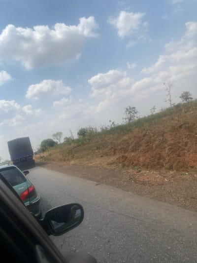 The scene depicts a roadside view in Ninzo West, Nigeria, under a bright blue sky with scattered white cumulus clouds. The perspective is from inside a vehicle, with the dark A-pillar and side mirror partially visible in the lower left foreground; a distorted reflection is present in the mirror. The road, an asphalt surface showing cracks and wear, extends into the distance. To the immediate right of the road is an unpaved shoulder of dark brown gravel leading to a sloped embankment of reddish-brown earth, sparsely covered with dry, light-brown vegetation and scattered trees, some appearing dormant or bare, alongside others with green foliage.
Ahead on the road, partially obscured by the foreground vehicle's window frame, is a light green Peugeot 406 sedan, identifiable by "406" text on its rear. Further ahead and to the left, a large dark-colored truck with a taut, dark canvas-like cover over its cargo area is also visible on the road. Both the sedan and the truck appear stationary or in slow-moving traffic. No discernible individuals are visible, and no specific actions or interactions are clearly depicted. The environment suggests a rural or semi-rural road segment.