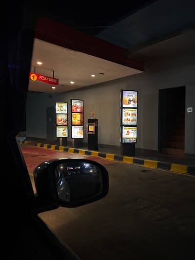 A nighttime view from inside a car at a McDonald's drive-thru in Kelapa Dua, Indonesia. The menu boards display food items and prices in Indonesian. A red sign indicates "Pesan Disini" (Order Here) with a number "1." The car's side mirror is visible, reflecting the scene behind. The drive-thru lane is marked with yellow and black striped barriers. The environment appears dimly lit.