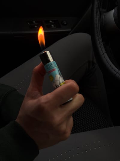 Inside a car, a hand holds a lit Clipper lighter with a cartoon design. The flame is directed towards a set of car window control buttons. The image is dark, with only the flame and lighter details visible. A steering wheel and car seat provide context. The angle suggests the photo was taken from the passenger seat.