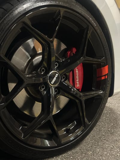 The image displays a close-up, angled view of the front wheel and tire of a white vehicle, located in Volketswil, Switzerland. The primary subject is a glossy black, multi-spoke alloy rim with a "SIGMA" branded center cap. Five lug nuts are visible around the center cap. A red brake caliper with white "SUPRA" lettering and two parallel red stripes is prominent behind the spokes. A metallic silver brake rotor is partially visible. The tire is black with visible tread and sidewall markings including "OUTSIDE / DOT T1D", "SuperEco", and partially visible "GOOD YEAR" branding. A portion of the white vehicle body is visible at the right edge. The wheel rests on a dark gray, speckled asphalt or concrete surface, suggesting an indoor or covered environment under artificial lighting. No individuals are present in the scene, and no active interactions or actions are depicted.