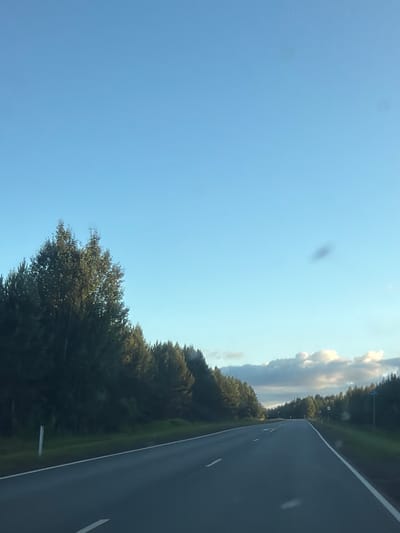 This is a media file of a road scene in Kamennyi Kliuch, Russia. The scene is captured from the perspective of someone traveling on a highway, indicated by the asphalt road with white lane markings on the right side of the frame. The road appears to be bordered by a dense line of green trees on the left. Above the trees, a clear blue sky dominates the upper portion of the image, with some scattered white clouds visible towards the horizon. The lighting suggests it is daytime, possibly late afternoon or early evening, given the angle of the light and the warm tones in the clouds. The overall mood is peaceful and serene, evoking a sense of travel and the natural beauty of the landscape. There are no visible people or text in the image.