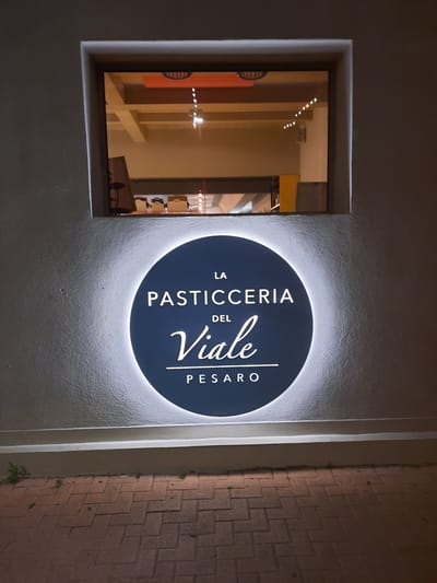 The image shows a circular, illuminated sign that reads "LA PASTICC.C.ERIA DEL Viale PESARO" in white lettering against a dark blue background. The sign is mounted on a textured, light-colored wall. Above the sign, a rectangular window reveals the interior of a building, with reflections of lights visible on the glass. The lower portion of the image shows a brick-paved walkway. The overall scene suggests the exterior of a pastry shop.