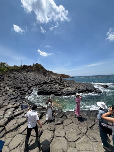 The image depicts the Gành Đá Đĩa (The Cliff of Stone Plates) in Phú Lương, Vietnam. The landscape is characterized by a dramatic formation of hexagonal basalt columns that descend into the sea. The sky is a clear blue with scattered white clouds. In the foreground, several people are present on the rocky shore. On the left, a man in a white shirt and black pants is walking away from the camera. To his right, a woman in a white dress and a man holding a baby are positioned near the water's edge. Further to the right, a woman in a pink floral dress and a white hat is facing the sea, and another person in a blue shirt and a white hat is seen from the back, holding a camera. The ocean exhibits moderate waves crashing against the rocks. A rope barrier with a sign is visible in the lower left quadrant of the image. The scene captures a tourist destination with natural geological formations and visitors engaging with the environment.