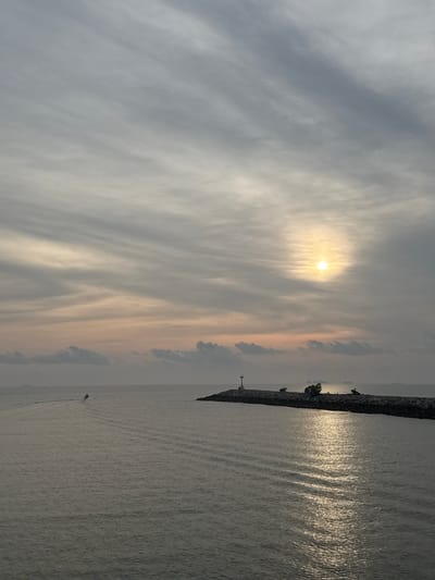 The image depicts a waterfront scene in Pak Nam Chumphon, Thailand at sunrise or sunset. The sun shines through a striated cloud cover, casting a golden glow. A long stone breakwater extends into the calm water. A small boat is visible on the water's surface.