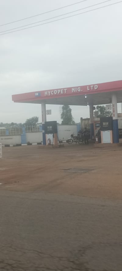 The image shows a Hycopet Nig. Ltd. gas station in Ladura Foron, Nigeria. The gas station has a red roof with white lettering displaying the company name. Several gas pumps are visible under the roof. A motorcycle is parked near one of the pumps, where people appear to be refueling. A fire extinguisher is mounted near a pump. In the background, there is a truck with a white container. The sky is overcast, and the ground appears to be dirt or gravel. Electrical power lines are visible in the upper part of the image.