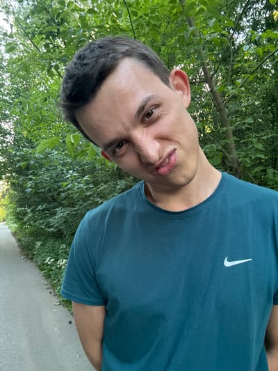 A young man, appearing to be in his late teens or early twenties, poses for a selfie outdoors. He is wearing a teal Nike t-shirt with the iconic swoosh logo visible on the left chest. The man has dark hair, brown eyes, and fair skin. His expression is a slight grimace. The background consists of lush green foliage, suggesting a natural setting such as a park or woodland. The lighting indicates it is daytime, likely late afternoon given the soft quality of light filtering through the leaves. The image is oriented sideways, with the man's head tilted to the right. The ground is a paved path, which appears to be a sidewalk or walking trail. There are no other people or significant objects in the frame besides the man and the natural surroundings. The location context provided is Шевлягино, Russia.