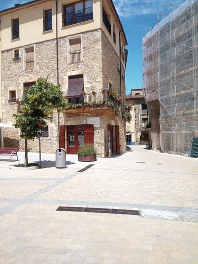 The image captures a street scene in the historic town of Besalú, Spain. The foreground is dominated by a paved square with light-colored, rectangular stone tiles. Two dark, rectangular drainage grates are visible in the pavement. To the left, a portion of a red metal bench is present, along with a small, round-leafed tree planted in a circular mulched area. A grey, perforated trash receptacle stands near the tree.

The main architectural feature is a building on the left with a stone facade on the lower levels and a cream-colored upper level. The stone is rough-hewn and varied in color, from light beige to gray. The building has multiple windows, some with dark metal railings and wooden shutters. A balcony with decorative railings and planters filled with flowers extends from the second story. A storefront with red wooden shutters and a glass door is visible at ground level.

To the right of the central building, a street or alleyway recedes into the background. This area features another building with a stone facade and a tiled roof. Further back, a taller building is covered in white scaffolding and a protective net, suggesting ongoing construction or renovation. The sky above is a clear blue with scattered white clouds. The overall impression is of a well-maintained, historic European town square on a bright, sunny day.