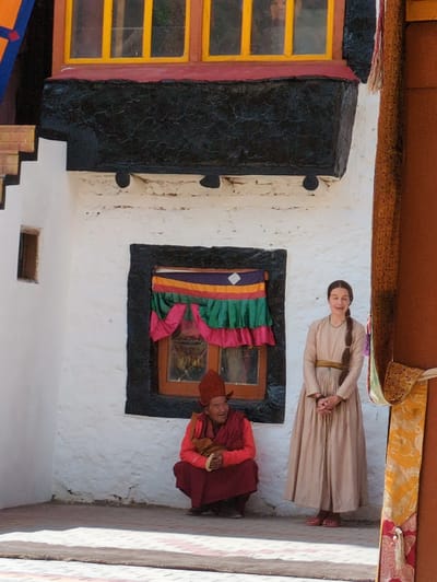 In the city of Pibiting, India, a scene unfolds in what appears to be a courtyard of a monastery or ancient building. The architecture is distinct, with a white stucco wall forming the backdrop. To the left, a section of the building features a dark, textured band below a window with yellow panes, above which are colored triangular elements. Further left, a small, dark rectangular window is set into the white wall.
Dominating the center is a window framed in black, adorned with a colorful curtain featuring horizontal bands of yellow, purple, green, and pink. To the left of this window, a person dressed in traditional red monk attire is seated in a squatting position. They are wearing a red cap and a red vest over a maroon robe, with a brown cloth held in their clasped hands. Their face is turned slightly upwards, and they appear to be smiling.
To the right of the window stands a young woman wearing a floor-length, pale beige dress with a darker belt. Her hair is braided and hangs down her back. She stands with her hands clasped in front of her. The right side of the image shows a large, reddish-brown structure, possibly a pillar or wall, with vibrant, patterned fabrics hanging alongside it, one predominantly yellow with intricate designs. The ground is paved with tiles, and a woven mat is visible in the foreground. The lighting suggests it is daytime, with shadows cast on the ground.