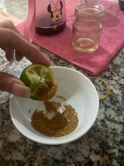 An overhead view reveals a human hand holding a half-cut green lime, its exposed flesh covered in a granular brown spice mixture, positioned above a white ceramic bowl containing a pile of the same mixture. The scene is located on a speckled grey granite countertop in a domestic setting within Đak Đoa, Vietnam. In the background, a pink water bottle featuring a "Minnie" character graphic rests on a pink fabric mat. Adjacent to it, a small, clear glass jar with a shallow residue of a clear yellowish liquid is also visible on the mat. A yellow rubber band lies on the countertop to the right of the bowl.
