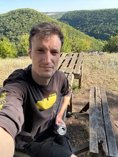 A young man is taking a selfie outdoors on a sunny day. He is sitting on a rustic wooden bench, holding a can of a beverage in his right hand. He is wearing a dark long-sleeved shirt with a yellow graphic on it, and dark pants. Behind him, a wooden picnic table is visible. The background features rolling, tree-covered hills and a clear blue sky. The area appears to be a natural landscape, possibly a park or a scenic viewpoint overlooking a valley. The location is identified as Velykozalissia, Ukraine.