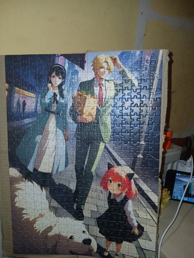 The image depicts a jigsaw puzzle featuring characters from the anime series Spy × Family. The puzzle is assembled and leaning against a wall with textured yellow paint. In the foreground of the puzzle, a fluffy white dog is partially visible. To the left, a woman with black hair and a blue dress stands with her hands clasped. In the center, a man with blond hair, wearing a suit and tie, carries a brown paper bag filled with groceries. To the right, a young girl with pink hair and black cat ears, dressed in a black dress and white shirt, is positioned in front of the man. The background of the puzzle shows a city street with architectural elements and blurred figures. The overall scene is likely captured indoors, as indicated by the indoor lighting and the presence of electrical cords and a phone or tablet on a surface to the right of the puzzle. There is no indication that this scene is located in Chutove, Ukraine; the image solely shows a puzzle and its immediate surroundings.