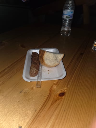 On a wooden surface, possibly a table, is a small white styrofoam tray containing a slice of bread and a sausage-like meat product. A small plastic utensil is also present on the tray. To the left of the tray is a partially visible plastic water bottle. The scene is dimly lit, with some areas in shadow. The location of the image is unspecified. The image does not contain any information about the location of Pripek, Bulgaria.