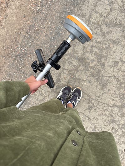 A first-person perspective shot shows a person standing on a grey concrete surface with small pebbles scattered around. The person is wearing a green corduroy jacket and pants, and black and white athletic shoes. They are holding a surveying instrument, which consists of a silver pole with a black handle, a device attached to the pole, and a circular grey and orange GPS unit at the top. The background shows a mix of grey concrete and gravelly dirt. The location is identified as Nevon, Russia.