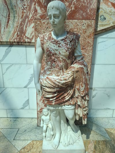 A full-body, eye-level, vertical shot shows a marble statue of a young, short-haired person in Roman-style armor, adorned with red and white marbled fabric. The statue stands on a white marble pedestal, partially obscuring a smaller, naked marble infant figure, which is seated on what appears to be a dolphin or similar marine creature. The main statue is positioned in front of a wall composed of large, polished marble slabs with striking red and white veining, some interspersed with green and lighter brown hues. To the left and right of the red-veined wall are sections of smooth, white marble tiles, creating a contrasting background. The floor is a mosaic of geometric marble tiles in shades of beige, brown, and grey, with a reflective surface. The lighting creates subtle shadows on the floor and around the statues. This scene is located within the grounds of the Trafford Centre, United Kingdom.