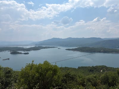 This media file shows a wide, scenic view of a large body of water with several tree-covered islands. In the foreground, dense green foliage from trees obscures the lower portion of the frame, suggesting the vantage point is from a hillside or elevated position. Beyond the immediate trees, the expanse of water dominates the middle ground. The water is calm and reflects the sky, with a subtle bluish-gray hue.
Several islands, varying in size and shape, are scattered across the water. These islands are densely covered in trees, creating a lush, green landscape. Some of these islands appear to be part of a larger landmass that curves around a section of the lake. In the distance, rolling hills and mountains form the backdrop, fading into a hazy blue. The sky above is partly cloudy, with white cumulus clouds scattered against a pale blue sky. The lighting suggests it is daytime, likely mid-morning or afternoon, with soft, diffused sunlight.
There are no visible people or specific activities taking place. The scene is serene and natural, evoking a sense of peacefulness and natural beauty. A thin black wire stretches across the middle of the frame, diagonally from the left to the right, a minor man-made element in an otherwise natural landscape. A tall, slender tower structure is faintly visible in the distance, rising from the treeline on one of the distant landforms. There is no visible text in the media file. The location context provided is Ponikvica, Montenegro, which aligns with the landscape's characteristics of a lake surrounded by hills and mountains.