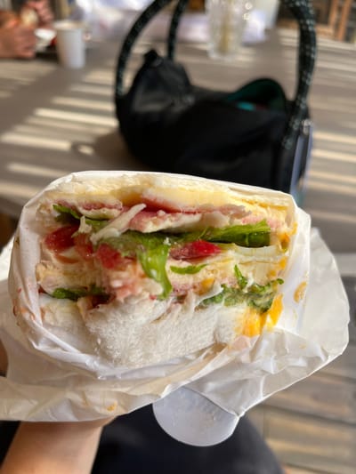 A large sandwich, partially wrapped in white paper, is held in someone's hand. Visible ingredients include white bread, ham, cheese, lettuce, tomato, and egg. The sandwich appears quite full, with filling spilling out from between the bread. A black bag with a green interior sits in the background to the left, partially obscured. A blurry outdoor setting with other people is faintly visible in the far background.