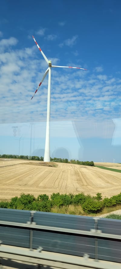 A single, tall, white wind turbine with red and white stripes on its blades stands prominently against a bright blue sky with scattered white clouds. The turbine's blades are angled, with one pointing upwards and to the right, and the other extending downwards and to the left. Below the turbine, a field of harvested, golden-brown wheat stretches across the landscape, showing clear rows of stubble. A line of green trees borders the far side of the field, with more distant wind turbines visible on the horizon. In the foreground, a dark gray, horizontal barrier or fence is visible, constructed of multiple slatted panels. The scene appears to be a rural or agricultural area. The reflection of another wind turbine and a person can be seen on a reflective surface in the middle ground, obscuring part of the view of the field and trees. The location is described as Wald am Arlberg, Austria.