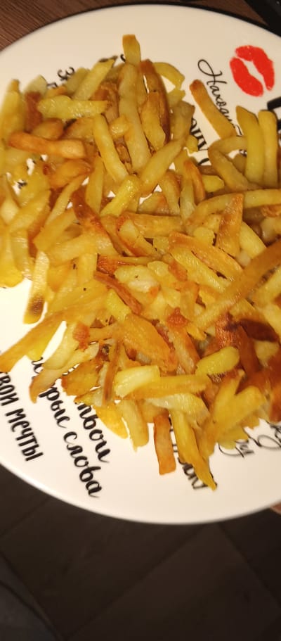 The image shows a white plate filled with french fries. The plate has black writing and a red lipstick kiss mark on it. The writing on the plate appears to be in Cyrillic script and includes the phrases "свои мечты" (svoi mechty - your dreams) and "добрые слова" (dobrye slova - kind words). The french fries are golden brown and appear to be freshly cooked. The background is dark and out of focus, suggesting the photo was taken indoors. The location is not specified in the image, therefore it cannot be determined if the scene is in Kondrovo, Russia.